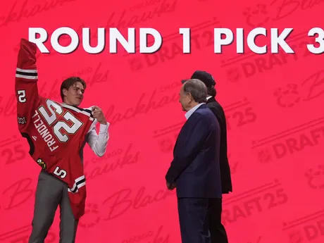 Anton Frondell (vľavo) si oblieka dres vedľa komisára NHL Garyho Bettmana (v popredí vpravo) a bývalého hokejistu Jeremyho Roenicka po tom, ako si ho v piatkovom drafte NHL 27. júna 2025 v Los Angeles vybral tím Chicago Blackhawks z tretieho miesta.