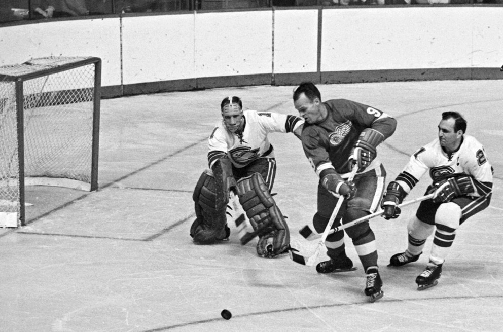 Na snímke z 26. októbra 1967 sa Gordie Howe (v strede) pokúša pretlačiť puk za chrbát brankára Californie Seals Charlieho Hodgea.