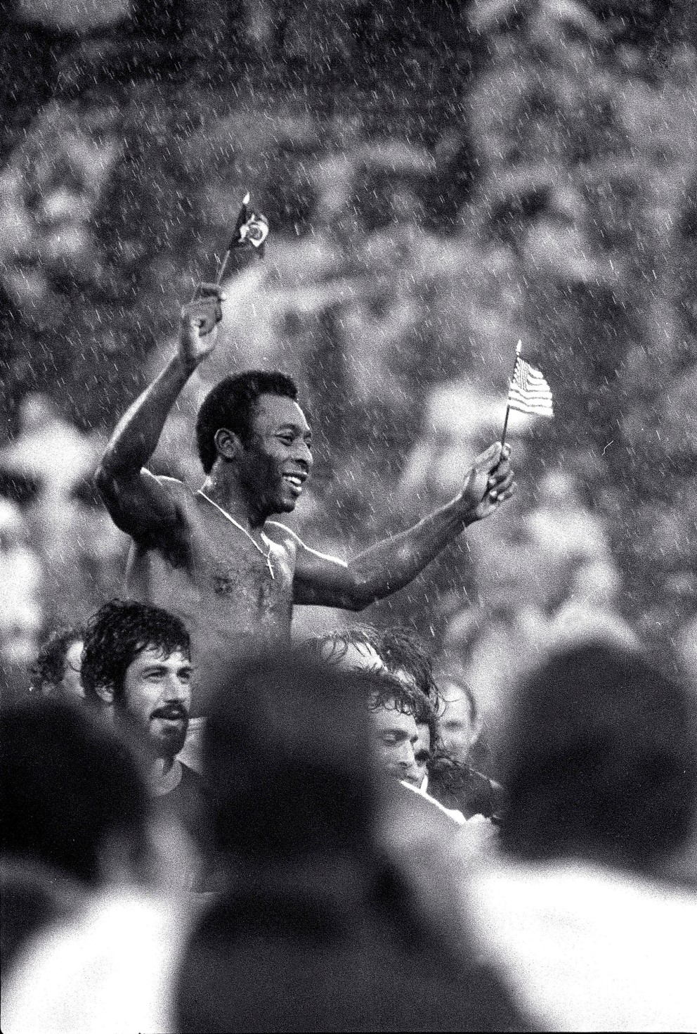 Futbalovú superhviezdu Pelého, mávajúcu vlajkami Brazílie a USA, odnášajú hráči oboch tímov z ihriska v silnom daždi na štadióne Giants v East Rutherford, N.J., po jeho poslednom zápase 1. októbra 1977.