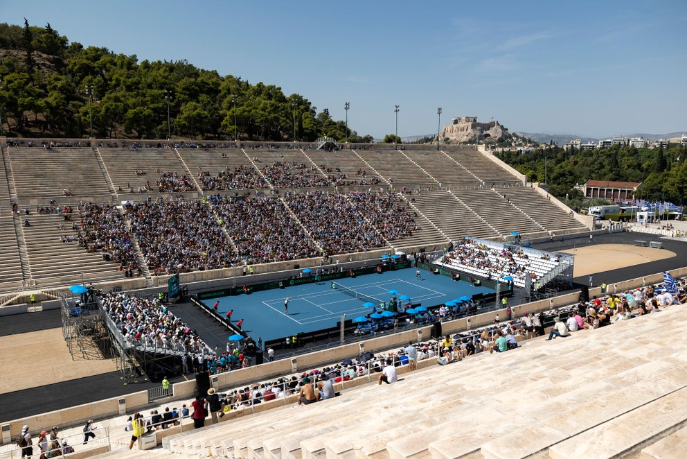 Pohľad na tenisový kurt na štadióne Panathenaic počas zápasu Tsitsipas - Klein. Na montovaných tribúnach boli biele sedačky, jedna z nich zostala poloprázdna.