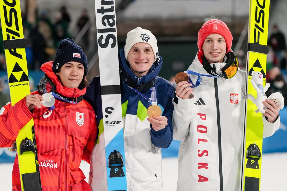Na snímke uprostred slovinský skokan na lyžiach Domen Prevc sa raduje po zisku zlata na veľkom mostíku na zimných olympijských hrách v meste Predazzo 14. februára 2026. Vľavo pózuje Japonec Ren Nikaidó so striebrom a vpravo Poliak Kacper Tomasiak s bronzom.